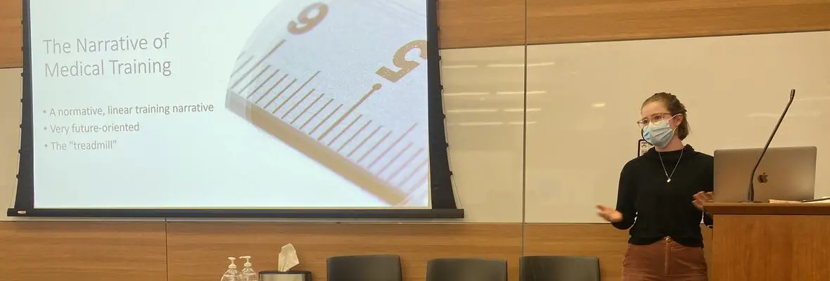 A woman in a black shirt and a surgical mask gestures with her hands as she speaks. To her left a projector screen reads 'The Narrative of Medical Training'