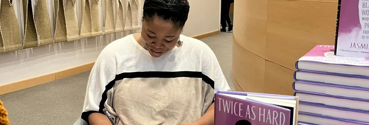 A woman in a white-and-tan shirt looks down as she writes. On the right side of the image, the top of a pink book cover that reads 'Twice as Hard' can be seen.